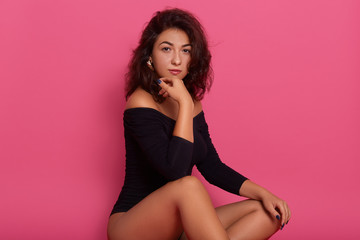 Brunette woman sitting on a floor, sensual female model wearing black body, keeping hand under chin, looking directly at camera isolated over rose background.