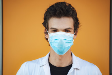 Close up portrait of young male doctor wearing face mask against yellow wall during COVID-19 outbreak