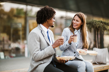 Smiling businessman and businesswoman with sandwiches sitting in front of the office building -...