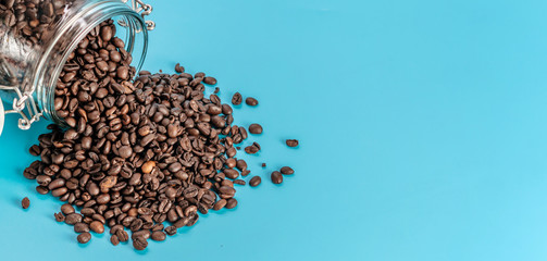 Aromatic roasted coffee beans spilled out of a glass jar on a blue background. Copy space