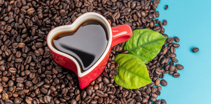 Red cup in the shape of a heart with freshly brewed black coffee and coffee beans scattered on a blue background