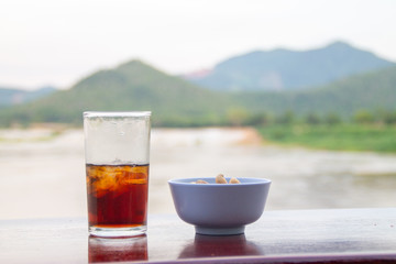 Cola in glass and bowl contain beans on table with mountain and river background