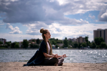 Fototapeta na wymiar A young girl with an afro braid in sportswear in a lotus position meditates on the beach. River and beautiful cloudy sky. Health and yoga concept.