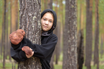 Beautiful girl in black sweater with hood hugs pine tree with large boletus in the forest.