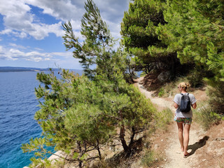 Young active feamle tourist wearing small backpack walking on coastal path among pine trees looking for remote cove to swim alone in peace on seaside in Croatia. Travel and adventure concept. - obrazy, fototapety, plakaty