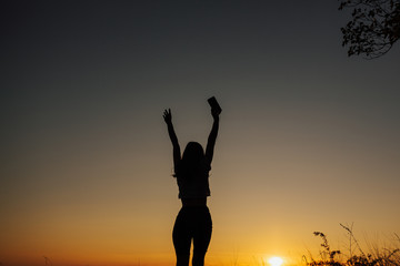 The girl at sunset. She stands back to the camera on the top of the mountain and looks at the sunset, welcomes the sun with her hands up. Girl listen the music.  Copy space.
