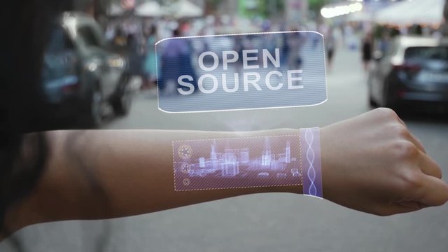 Female hand activates conceptual HUD holograms on smart bracelet with text Open source. Unrecognizable woman with holographic technology on a city street background
