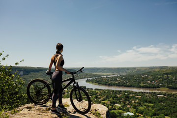 Attractive sporty girl standing on the rock on mountain against the backdrop river, town and blue sky. Young woman with bicycle riding in the mountains. 
