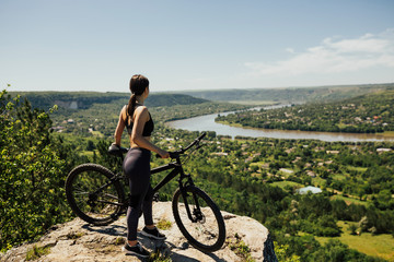 Fototapeta na wymiar Sporty girl with bicycle on the top of rock above the river and town. She enjoying the views of the mountain and the river. Copy space.