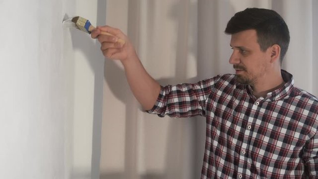 Portrait of man house painter is painting wall in grey using brush doing renovation at home, side view. DIY project. Young guy professional decorator at work in object at apartment, house.