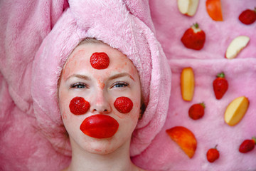 A young girl with a pink towel on her head lies with a mask of strawberries on her face. Nearby pieces of fruit