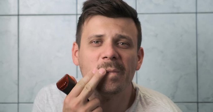 Young man is shaving beard and moustache using electric razor, front view. He is looking at camera and smiling in slow motion. Daily morning routine. Hygiene procedure.