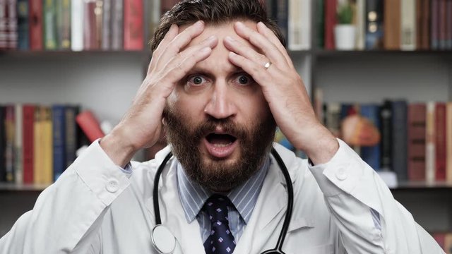 Horror doctor. Frightened excited male doctor in white coat and stethoscope in office looks at camera and covers his face with his hands