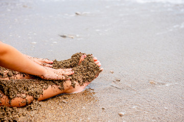 child plays on the beach in the sand. Baby hands and feet in the sand on the beach close-up. A child builds a sand castle. A child on the seashore. By the ocean. Dirty sand by the sea