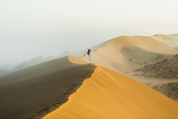 Fototapeta na wymiar Woman walking on top of huge sand dune in Morocco Sahara desert. Beautiful warm sun light and mist in morning. Freedom concept.
