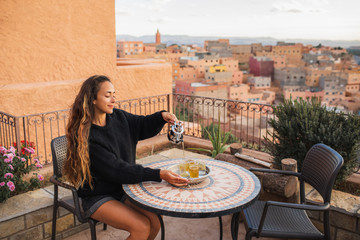 Woman pouring traditional moroccan mint tea on terrace with amazing view of old arab town Boumalne in Morocco at sunset. Round vintage table. - 371622723