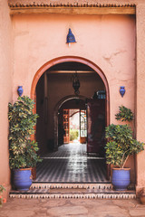 Fototapeta na wymiar Traditional moroccan style in architecture with red terracotta wall. Arch entrance, blue lanterns and clay flower pots with plants. Perspective inside.