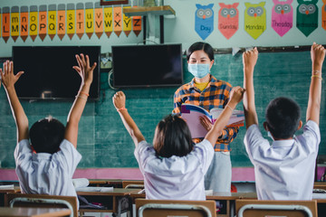 Obraz premium Young Asian female teachers wearing a medical face and students in a rural Thai village school are learning, grinding and raising their hands to answer teacher questions.