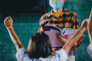 Young Asian female teachers wearing a medical face and students in a rural Thai village school are learning, grinding and raising their hands to answer teacher questions.