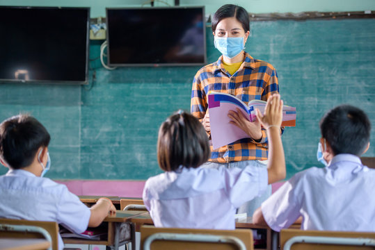 Young Asian female teachers wearing a medical face and students in a rural Thai village school are learning, grinding and raising their hands to answer teacher questions.