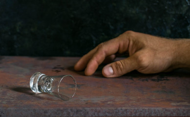 lying empty glass of vodka or alcoholic drink, hand drunk in the background, concept of alcoholism and alcohol abuse, defocused, selective focus, close-up, gray table, dark background