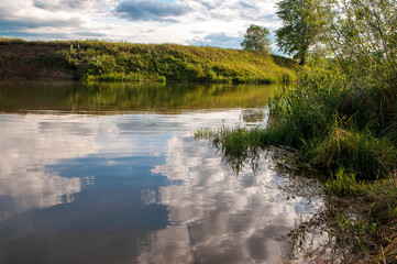 a narrow river with a mirror of blue sky with clouds, with a high Bank on the other side