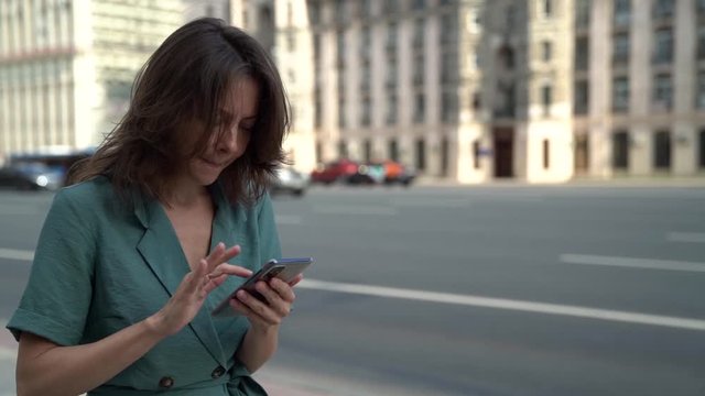 A cheerful adult woman walks in the city on a summer day along the highway with cars against the background of beautiful architecture, slow traffic. She looks at her smartphone.
