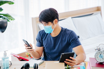 Serious man wear mask making note and checking on mobile phone while sitting on Desk in the Bedroom. COPY SPACE. Trading. WORK FROM HOME Concept.