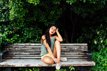 Latin young girl with long black hair and blue dress, sitting on wooden bench among greenery, looking at camera, romantic concept