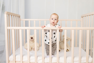 a baby of 8 months stands in a crib with toys in pajamas in a bright children's room after sleeping...