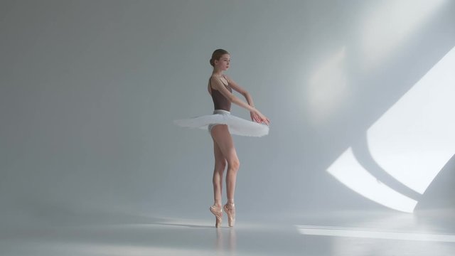 A professional ballerina in a white tutu dances in a large training hall. Girl does dance steps in the stage costume. Shot on a white background in the spacious and brightly lit studio.