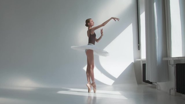 Young ballerina training in the studio standing by the window. Flexible girl on a white background in bright light.