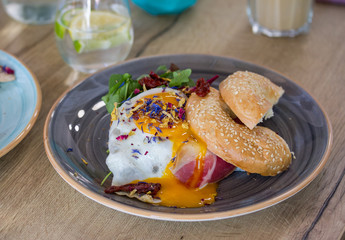 Gourmet breakfast bagels with a sunny side up egg, ham and edible flower decoration on a grey plate on wooden table in a restaurant