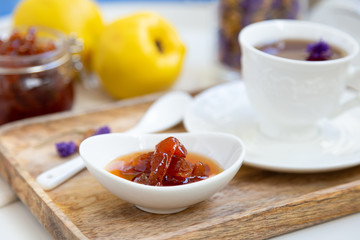 Homemade quince jam in a porcelain bowl with a cup of tea on a wooden tray. In the background is a glass jar with jam and quince.