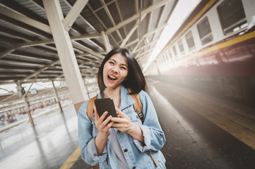 Fototapeta premium Happy smiling Asian woman tourist travel with backpacker holding smartphone at train station platform