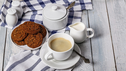 Coffee in a white cup, cream, sugar bowl and oatmeal cookies on a wooden table. Copy space