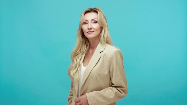 Angry middle aged blond woman in business suit touching nose and pointing finger to camera, showing liar gesture, suspecting and blaming in lying. Indoor studio shot isolated on blue background