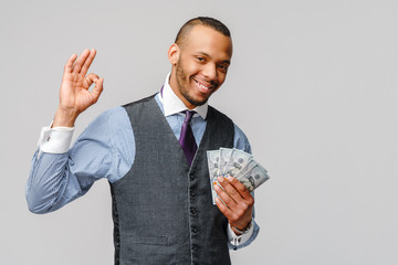 Portrait of excited young african american man holding cash money and showing OK sign over light...
