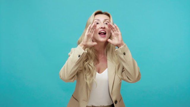 Smiling positive middle aged woman loudly shouting holding hands near mouth, calling about announcement, inviting. Indoor studio shot isolated on blue background