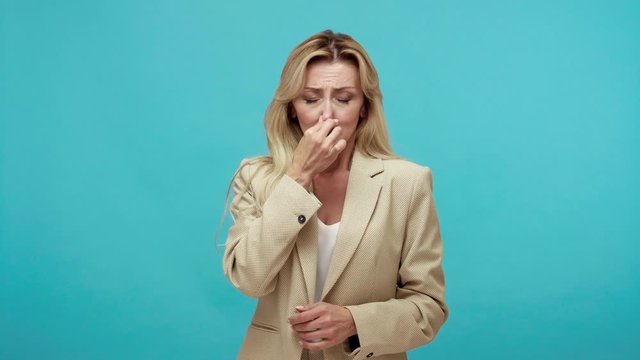 Intelligent middle aged blond woman in business style suit closing her nose with fingers feeling unpleasant disgusted smell. Indoor studio shot isolated on blue background