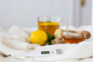 Selective focus on thermometer on light background. Cup of tea, lemon, mint, ginger root and honey for colds or fever on light background. Alternative medicine and home flu treatment