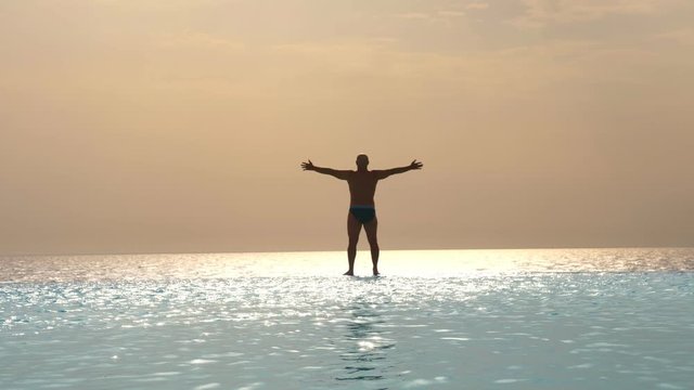 dark silhouette, man stands with arms outstretched to sides and relaxing on edge of outdoor infinity pool with panoramic sea view, at sunrise. inspiration, travel and vacation concept