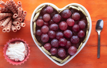 Close-up view of the white heart-shaped plate filled with ripe red grapes, accompanied by cream and biscuits. Wooden background