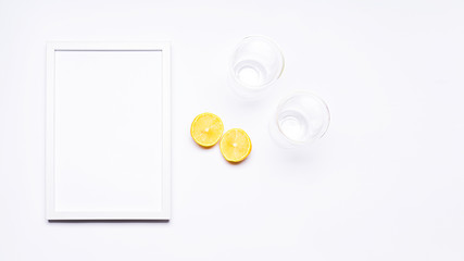 Summer composition. White wooden frame with empty space for text. Next to it, there is a lemon cut in half and there are two glasses. Everything is arranged on a white background. Flat top view.