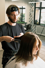 Professional Hairstylist with beard  in a black t-shirt and bandana who making hair dress for young white woman. Concept of barber and hairdresser industry.