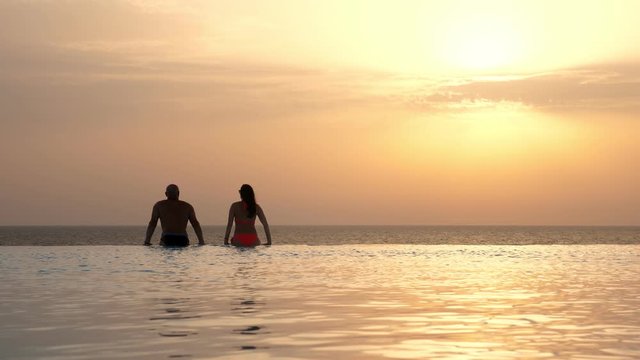 back view, silhouettes, couple in love, man and woman, relaxing in water, on edge of outdoor infinity pool with panoramic sea view, at sunrise. honeymoon, travel and vacation concept