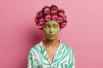 Funny ethnic young woman makes grimace, crosses eyes, applies hair rollers, makes hairstyle for special day, wears green moisturizing mask on face, dressed in domestic clothes, poses in studio