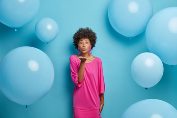Fototapeta premium Romantic Afro American woman sends air kiss, keeps palm forward camera, expresses love and affection, wears elegant pink dress, poses against blue background with inflated balloons. Party time