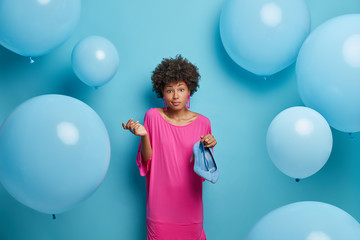 Fototapeta premium Indoor shot of hesitant Afro American woman holds blue shoes, decides what to wear on special occasion, stands unaware, shrugs shoulders, dressed in festive clothes, poses over decorated background