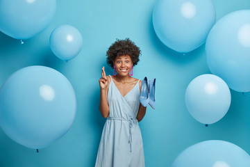 Hopeful young Afro American woman crosses fingers, makes wish, wears high heel shoes and dress, dresses for party, stands indoor, blue background, inflated balloons around. Holiday preparation